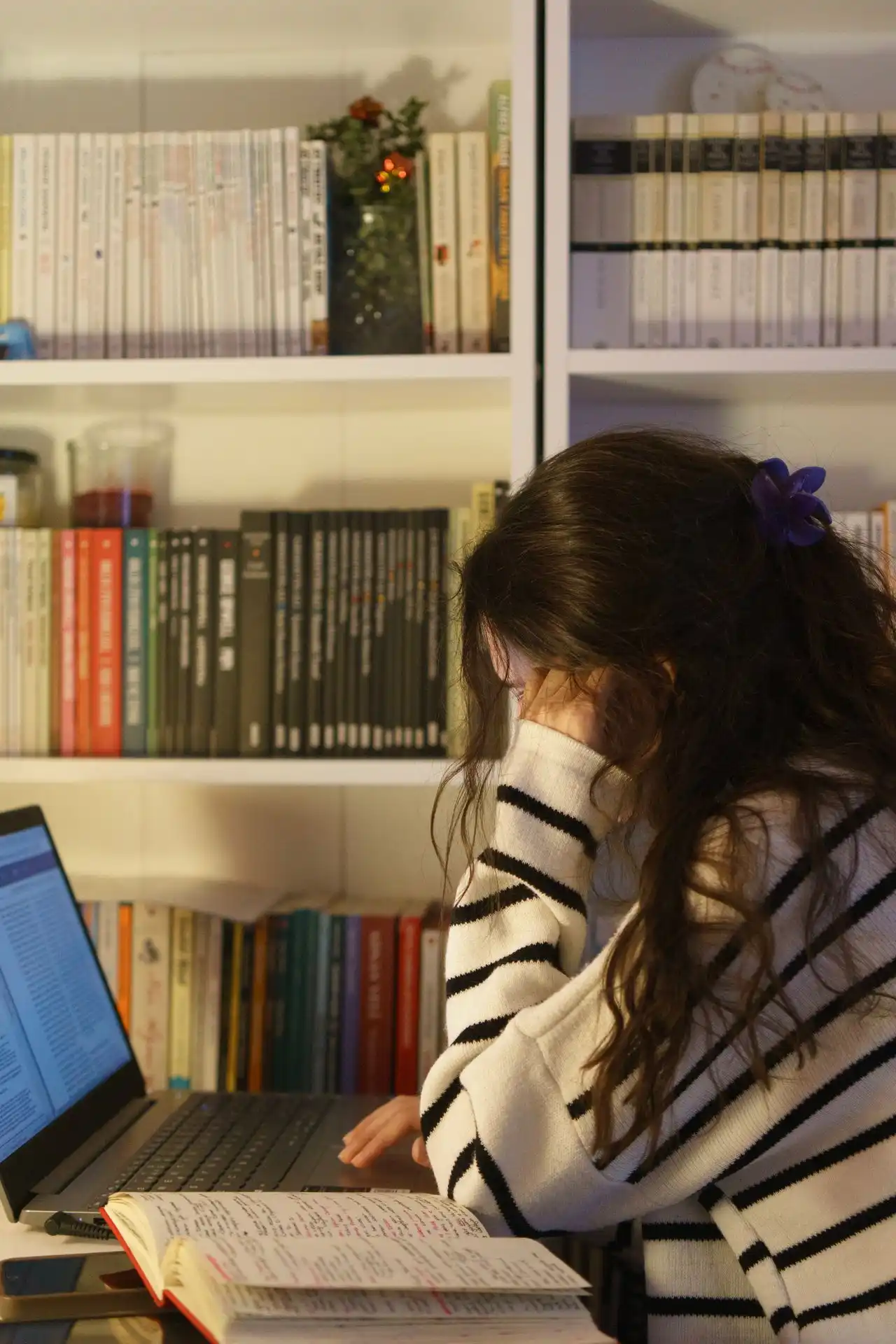Estudiante consultando su laptop y un cuaderno frente a una biblioteca